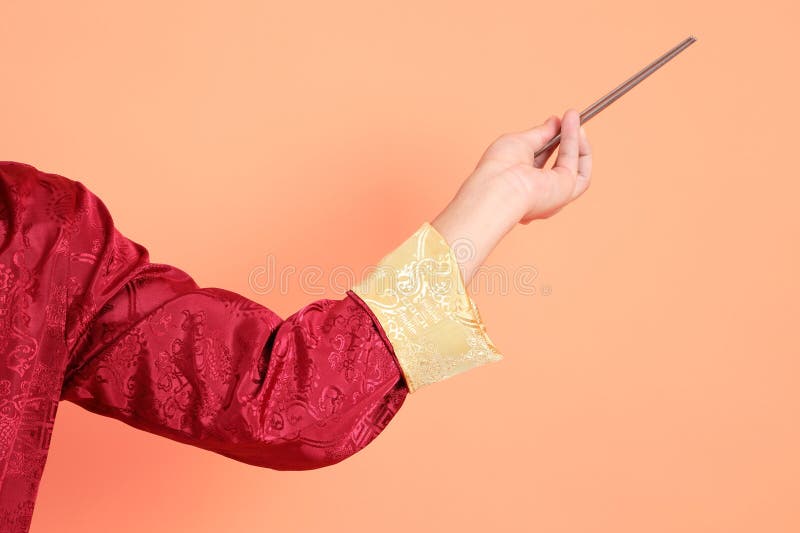 Happy Chinese new year. Hand of asian chinese senior man wearing red traditional cheongsam qipao or changshan dress with gesture of hand holding silver chopsticks isolated on orange background. Happy Chinese new year. Hand of asian chinese senior man wearing red traditional cheongsam qipao or changshan dress with gesture of hand holding silver chopsticks isolated on orange background