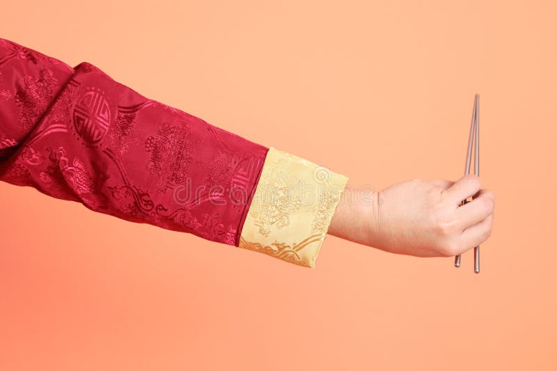 Happy Chinese new year. Hand of asian chinese senior man wearing red traditional cheongsam qipao or changshan dress with gesture of hand holding silver chopsticks isolated on orange background. Happy Chinese new year. Hand of asian chinese senior man wearing red traditional cheongsam qipao or changshan dress with gesture of hand holding silver chopsticks isolated on orange background