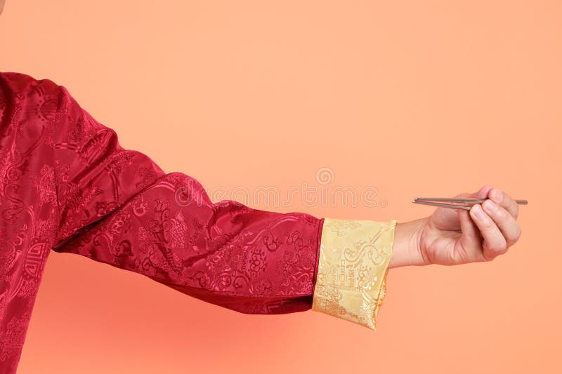 Happy Chinese new year. Hand of asian chinese senior man wearing red traditional cheongsam qipao or changshan dress with gesture of hand holding silver chopsticks isolated on orange background. Happy Chinese new year. Hand of asian chinese senior man wearing red traditional cheongsam qipao or changshan dress with gesture of hand holding silver chopsticks isolated on orange background