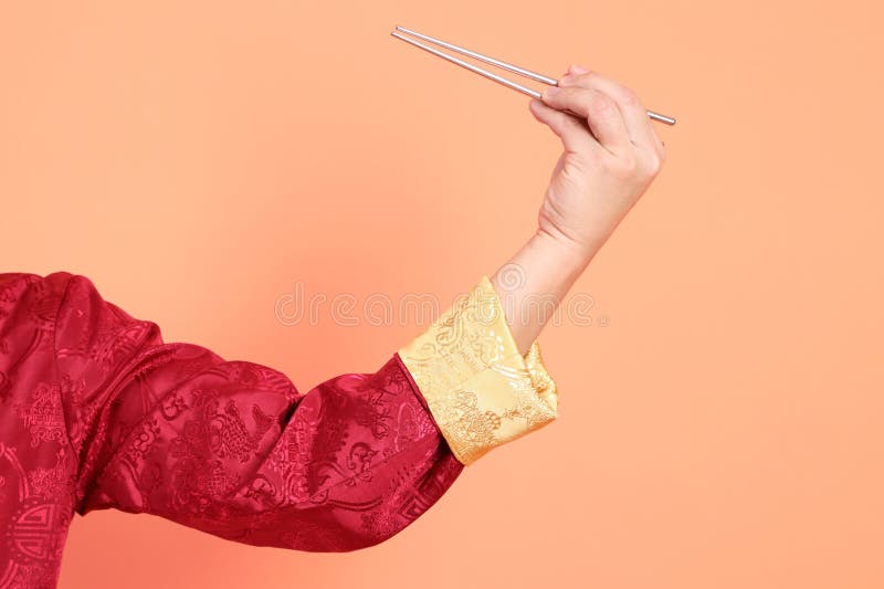 Happy Chinese new year. Hand of asian chinese senior man wearing red traditional cheongsam qipao or changshan dress with gesture of hand holding silver chopsticks isolated on orange background. Happy Chinese new year. Hand of asian chinese senior man wearing red traditional cheongsam qipao or changshan dress with gesture of hand holding silver chopsticks isolated on orange background