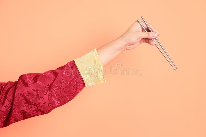 Happy Chinese new year. Hand of asian chinese senior man wearing red traditional cheongsam qipao or changshan dress with gesture of hand holding silver chopsticks isolated on orange background. Happy Chinese new year. Hand of asian chinese senior man wearing red traditional cheongsam qipao or changshan dress with gesture of hand holding silver chopsticks isolated on orange background