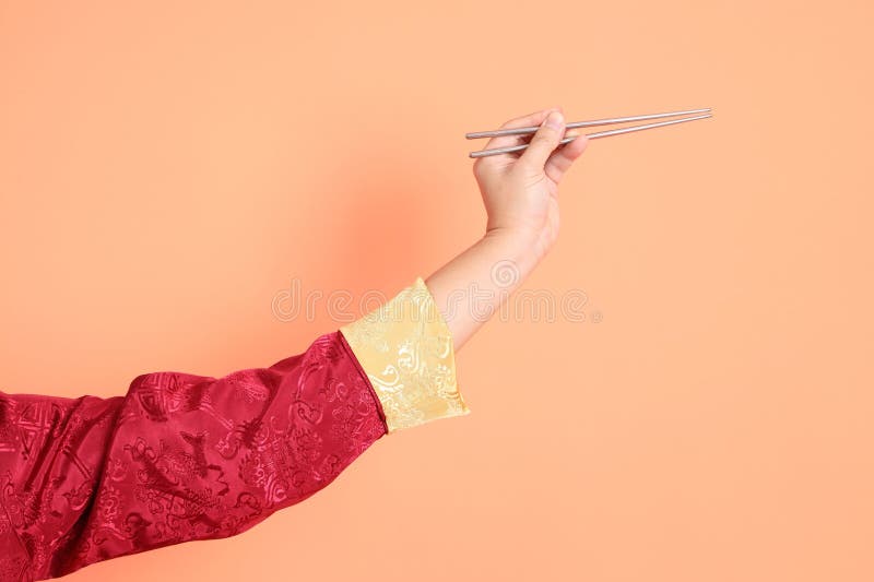 Happy Chinese new year. Hand of asian chinese senior man wearing red traditional cheongsam qipao or changshan dress with gesture of hand holding silver chopsticks isolated on orange background. Happy Chinese new year. Hand of asian chinese senior man wearing red traditional cheongsam qipao or changshan dress with gesture of hand holding silver chopsticks isolated on orange background