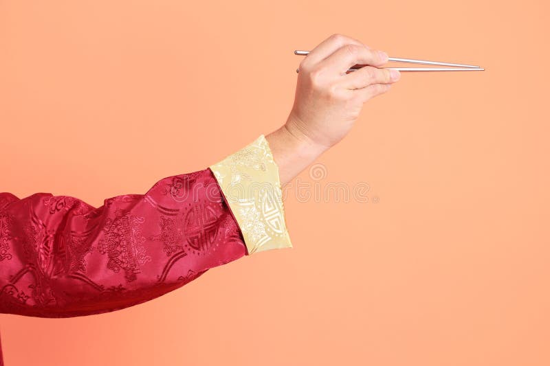 Happy Chinese new year. Hand of asian chinese senior man wearing red traditional cheongsam qipao or changshan dress with gesture of hand holding silver chopsticks isolated on orange background. Happy Chinese new year. Hand of asian chinese senior man wearing red traditional cheongsam qipao or changshan dress with gesture of hand holding silver chopsticks isolated on orange background