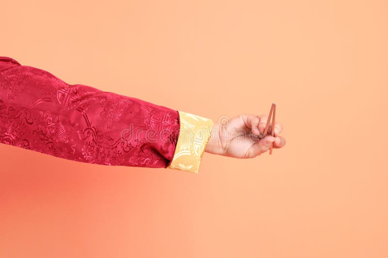 Happy Chinese new year. Hand of asian chinese senior man wearing red traditional cheongsam qipao or changshan dress with gesture of hand holding silver chopsticks isolated on orange background. Happy Chinese new year. Hand of asian chinese senior man wearing red traditional cheongsam qipao or changshan dress with gesture of hand holding silver chopsticks isolated on orange background