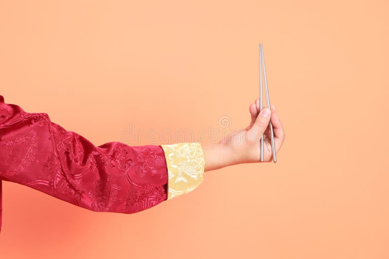 Happy Chinese new year. Hand of asian chinese senior man wearing red traditional cheongsam qipao or changshan dress with gesture of hand holding silver chopsticks isolated on orange background. Happy Chinese new year. Hand of asian chinese senior man wearing red traditional cheongsam qipao or changshan dress with gesture of hand holding silver chopsticks isolated on orange background