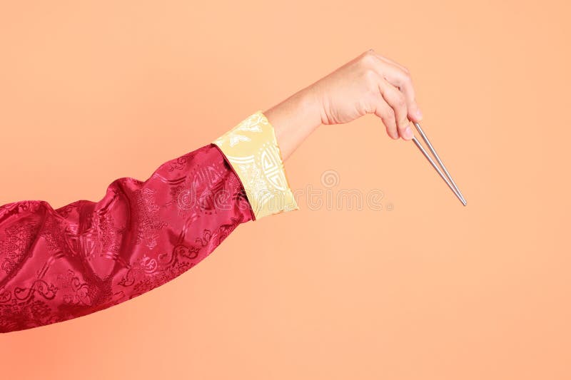 Happy Chinese new year. Hand of asian chinese senior man wearing red traditional cheongsam qipao or changshan dress with gesture of hand holding silver chopsticks isolated on orange background. Happy Chinese new year. Hand of asian chinese senior man wearing red traditional cheongsam qipao or changshan dress with gesture of hand holding silver chopsticks isolated on orange background