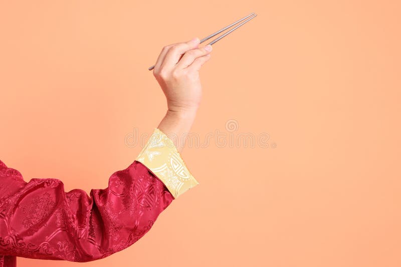 Happy Chinese new year. Hand of asian chinese senior man wearing red traditional cheongsam qipao or changshan dress with gesture of hand holding silver chopsticks isolated on orange background. Happy Chinese new year. Hand of asian chinese senior man wearing red traditional cheongsam qipao or changshan dress with gesture of hand holding silver chopsticks isolated on orange background