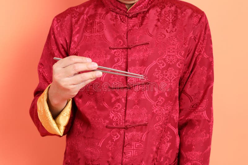 Happy Chinese new year. Hand of asian chinese senior man wearing red traditional cheongsam qipao or changshan dress with gesture of hand holding silver chopsticks isolated on orange background. Happy Chinese new year. Hand of asian chinese senior man wearing red traditional cheongsam qipao or changshan dress with gesture of hand holding silver chopsticks isolated on orange background