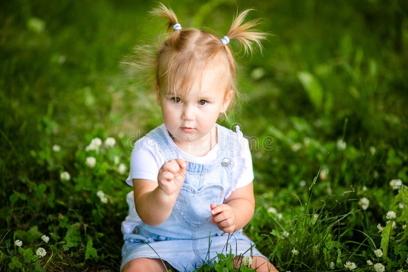 Feliz Bebê Loira Com Duas Pequenas Tranças Em Camiseta Branca E Um Kit De  Calças Jeans Sentado Sobre O Fundo Da Grama Verde Foto de Stock - Imagem de  pouco, inocência: 143643548
