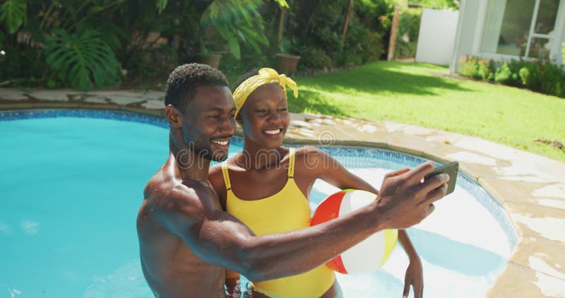 Feliz casal africano-americano em pé na piscina tomando selfie e sorrindo no jardim ensolarado