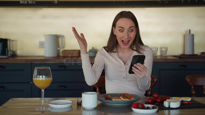 Feliz e linda jovem na cozinha, com seu telefone jogando on-line ganha prêmio. ele mostra um vitorioso