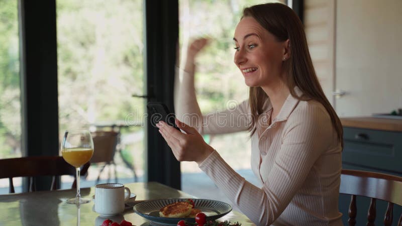 Feliz e linda jovem na cozinha, com seu telefone jogando on-line ganha prêmio. ele mostra um vitorioso