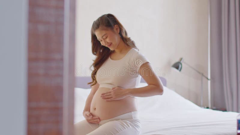 Feliz embarazada asiática sentada en la cama sosteniendo y acariciando su gran barriga en casa embarazo de una joven que disfruta almacen de metraje de vídeo