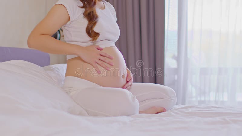 Feliz embarazada asiática sentada en la cama sosteniendo y acariciando su gran barriga en casa embarazo de una joven que disfruta metrajes