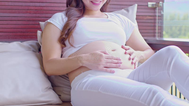 Feliz embarazada asiática sentada en la cama sosteniendo y acariciando su gran barriga en casa embarazo de una joven que disfruta almacen de video