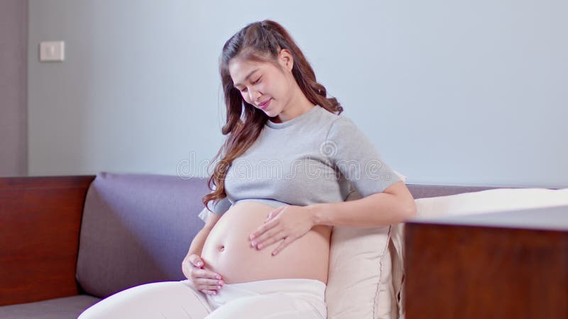Feliz embarazada asiática sentada en la cama sosteniendo y acariciando su gran barriga en casa embarazo de una joven que disfruta almacen de metraje de vídeo