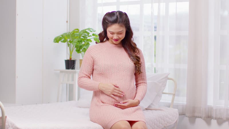 Feliz embarazada asiática sentada en la cama sosteniendo y acariciando su gran barriga en el embarazo de su casa de una joven disf metrajes