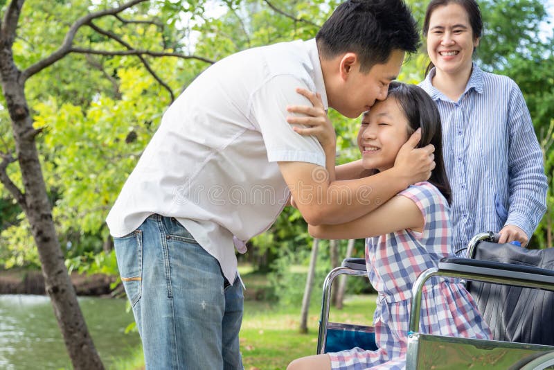 Feliz famÃlia asiática, linda filha doente em cadeira de rodas enquanto seu pai se abraçava, beijando na testa ela e a mãe sor imagem de stock Feliz famÃlia asiática, linda filha doente em cadeira de rodas enquanto seu pai se abraçava, beijando na testa ela e a mãe sor imagem de stock