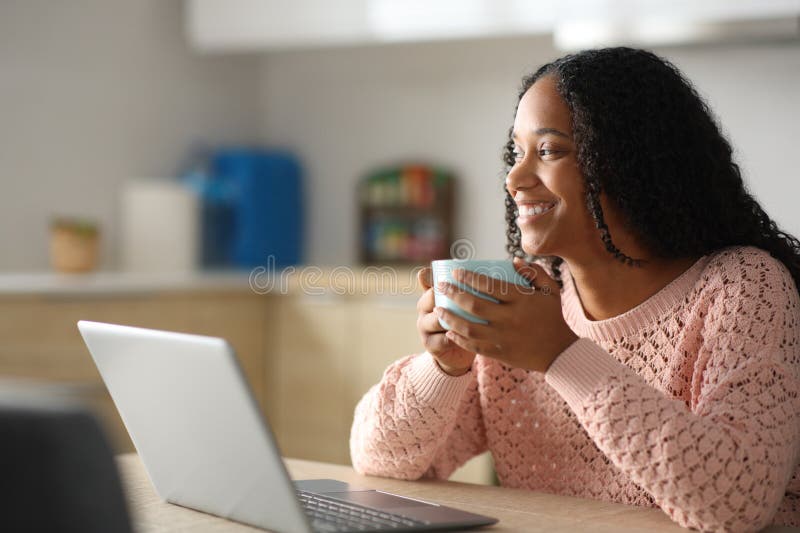 Feliz mujer negra con un portátil tomando café en la cocina fotos de archivo