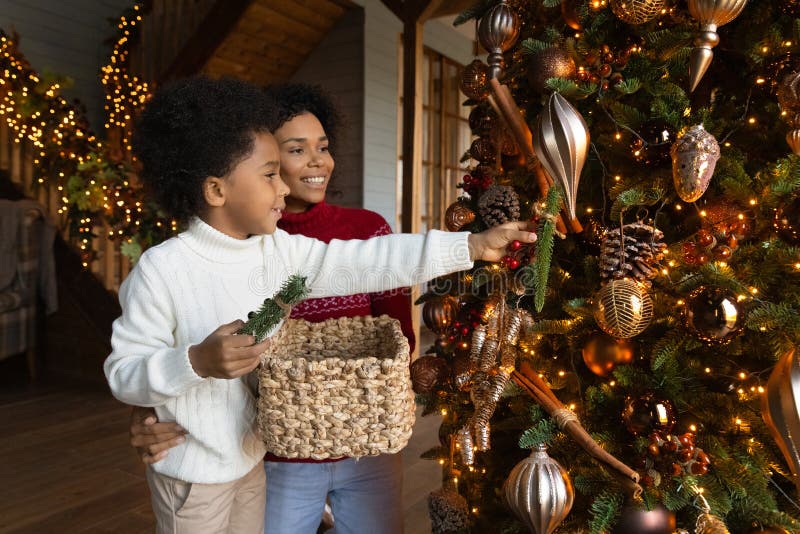 Happy African American mother and little son decorating Christmas tree with baubles, holding basket with toys, family wearing sweaters preparing home for New Year party, enjoying winter holiday. Happy African American mother and little son decorating Christmas tree with baubles, holding basket with toys, family wearing sweaters preparing home for New Year party, enjoying winter holiday