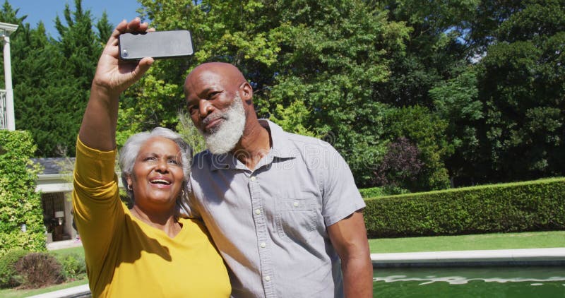 Feliz senior afroamericana pareja usando smartphone tomando selfie por piscina en jardín soleado