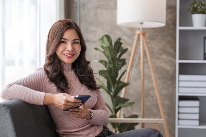 Feliz y bella mujer asiática sentada en el sofá con la mano sosteniendo un teléfono móvil charlando con amigos y jugando a los med fotos de archivo libres de regalías