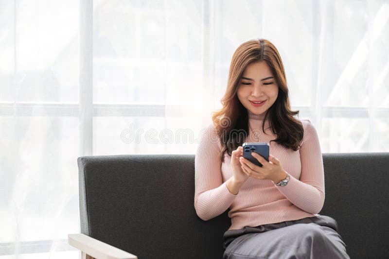 Feliz y bella mujer asiática sentada en el sofá con la mano sosteniendo un teléfono móvil charlando con amigos y jugando a los med foto de archivo