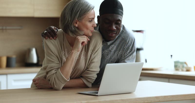 Feliz y diversa pareja milenaria de citas usando laptop en la cocina de casa