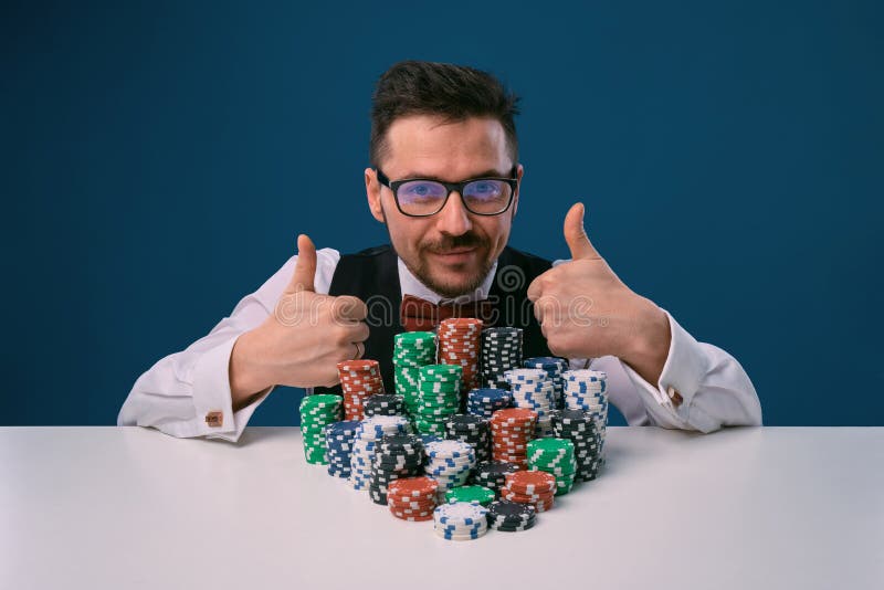 Fellow in glasses, black vest and shirt sitting at white table with stacks of chips on it, posing on blue background stock image