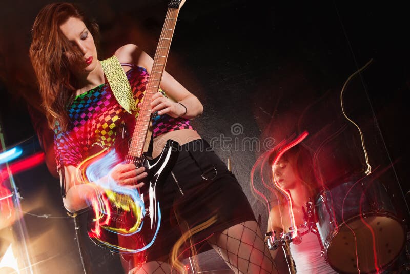 Photo of a young beautiful women playing guitar with drummer in the background. Motion blur and light trails from slow shutter speed and strobes. Space for text. Photo of a young beautiful women playing guitar with drummer in the background. Motion blur and light trails from slow shutter speed and strobes. Space for text.