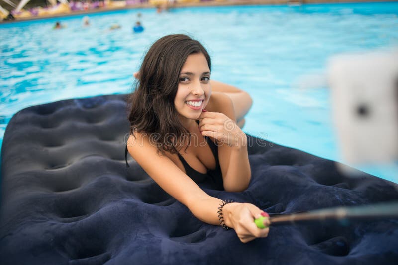 Attractive female in bikini smiling and makes selfie photo on the phone with selfie stick on a mattress in the pool at the resort.