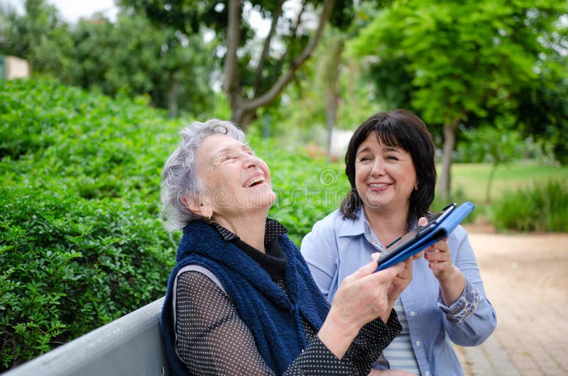 Senior adult woman and a carer laugh holding a tablet in their hands while sitting on a park bench royalty free stock photography Senior adult woman and a carer laugh holding a tablet in their hands while sitting on a park bench royalty free stock photography