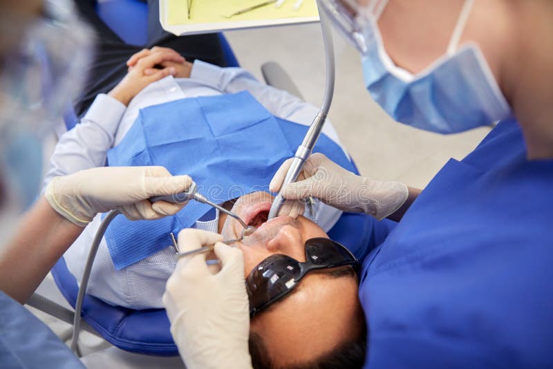 People, medicine, stomatology and health care concept - female dentist and assistant with dental mirror, drill and air water gun spray treating male patient teeth at dental clinic. People, medicine, stomatology and health care concept - female dentist and assistant with dental mirror, drill and air water gun spray treating male patient teeth at dental clinic