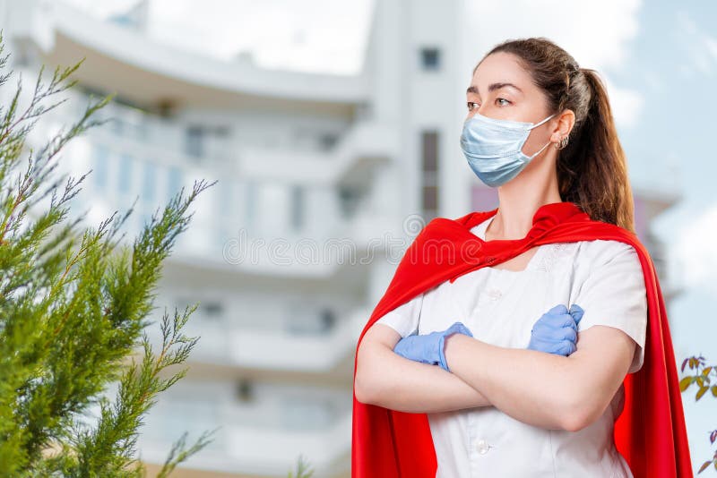 A female doctor wearing a surgical mask and rubber gloves poses with her arms crossed and a super hero&#x27;s cape on her back