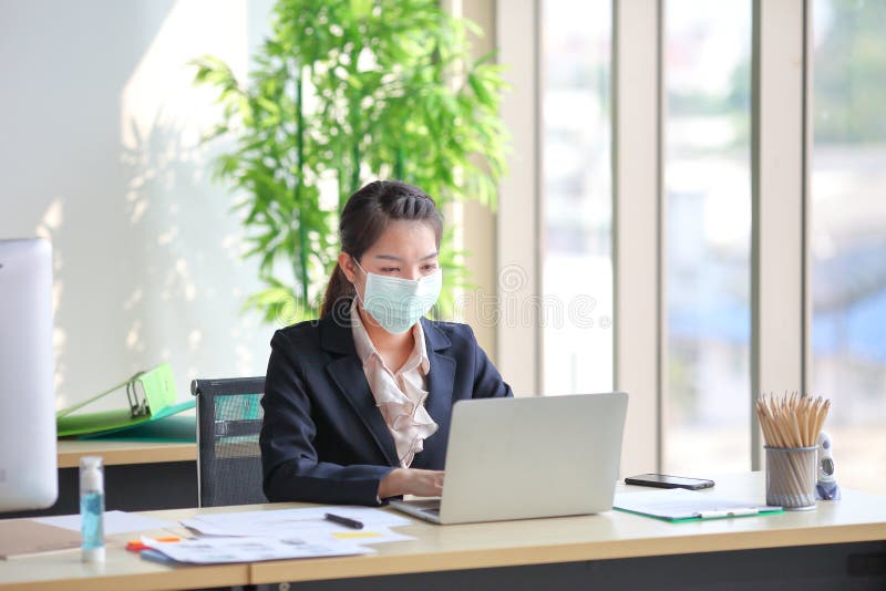 Female employee wearing medical face mask while working alone because of social distancing policy in the business office