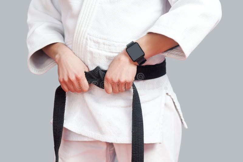 Female fighter torso poses in white kimono, smartwatch and sportive strong arms are closeup black belt. sport and technology healthcare concept. Indoor studio shot, on grey background