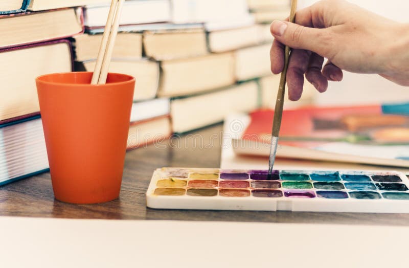 Female hand draws with a brush and watercolor paint on a sheet of paper on wooden table with stack of books background royalty free stock photography Female hand draws with a brush and watercolor paint on a sheet of paper on wooden table with stack of books background royalty free stock photography