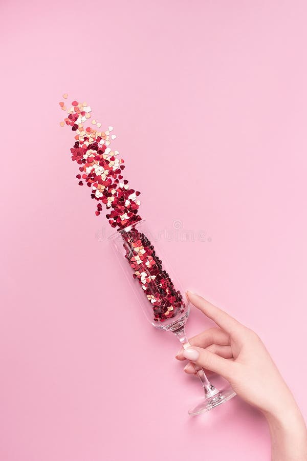 Female hand holding champagne glass with vibrant red heart-shaped confetti poured out of it on pink background. Flat lay