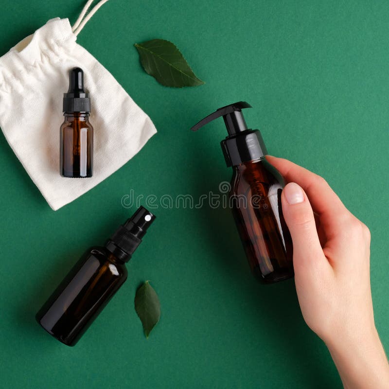 Female hand holding pump amber glass bottle over green background with spray bottle and essential oil dropper bottle. SPA natural