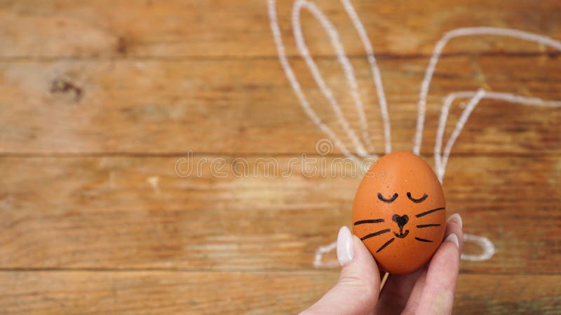 A female hand holds an egg with a cute face. The ears are drawn with chalk