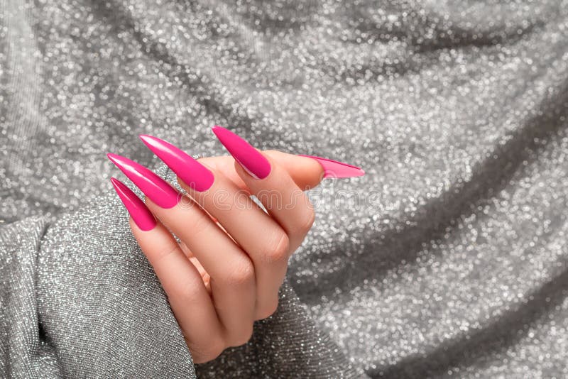 Female hand with pink stiletto nail design. Long nail polish manicure. Woman hand on glitter silver fabric background