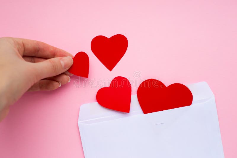 Female hand puts paper red hearts in a white envelope. Love letter. Love message. Valentine`s day concept. Happy Valentine's day greeting card. Female hand puts paper red hearts in a white envelope. Love letter. Love message. Valentine`s day concept. Happy Valentine's day greeting card.