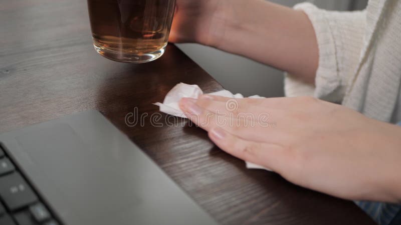 Female hand wipes spilled liquid on wooden table with paper napkin, glass of tea in his right hand. Close-up Slow Motion