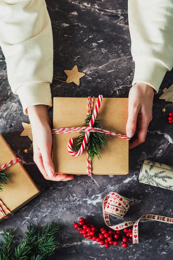 Female hands holding Christmas gift box decorated evergreen branch, striped ribbon, candy cane on stone background. Merry