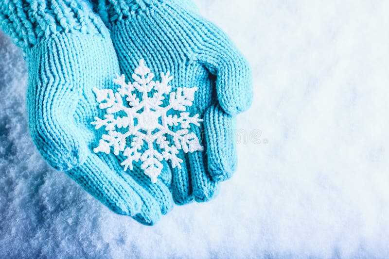 Female hands in light teal knitted mittens with sparkling wonderful snowflake on a white snow background. Winter Christmas concept