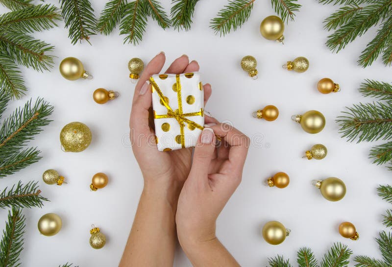 Female hands opens a small gift box on white background with golden christmas balls and green fir branches. Winter flat