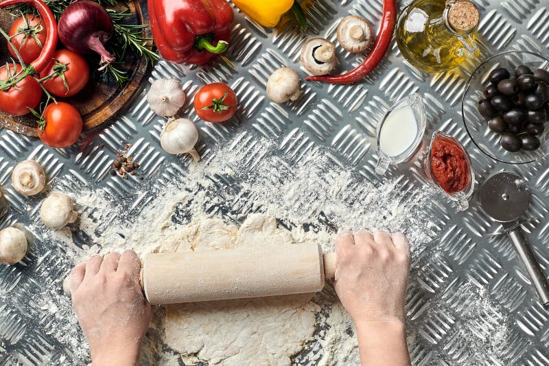 Female hands roll out dough on metal background, close up. Chef makes dough. Table with vegetables and kitchenware stock photo Female hands roll out dough on metal background, close up. Chef makes dough. Table with vegetables and kitchenware stock photo