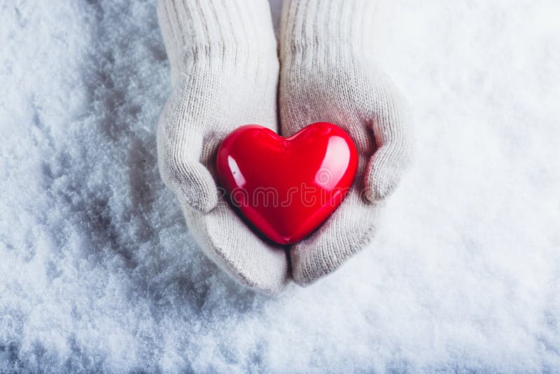 Female hands in white knitted mittens with a glossy red heart on a snow background. Love and St. Valentine concept.