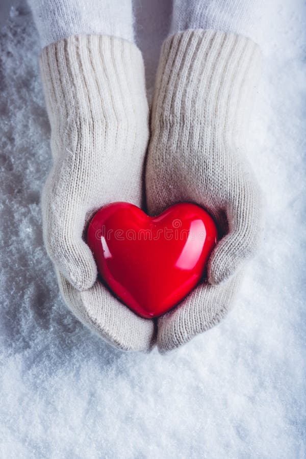 Female hands in white knitted mittens with a glossy red heart on a snow background. Love and St. Valentine concept.