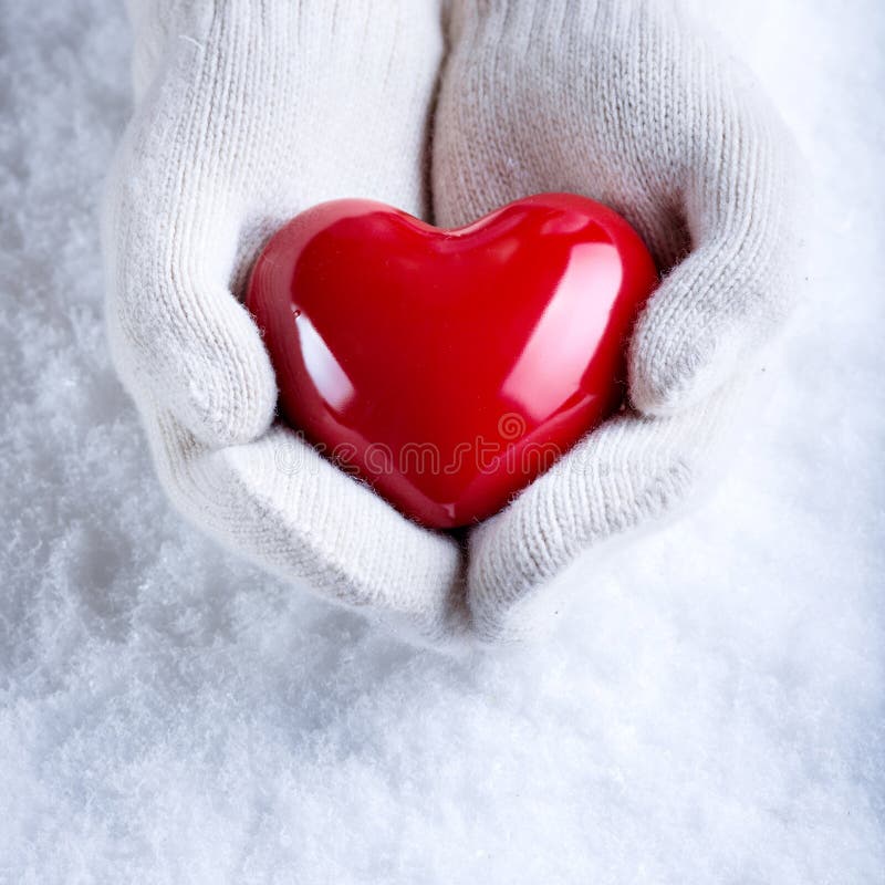 Female hands in white knitted mittens with a glossy red heart on a snow winter background. Love and St. Valentine cozy concept